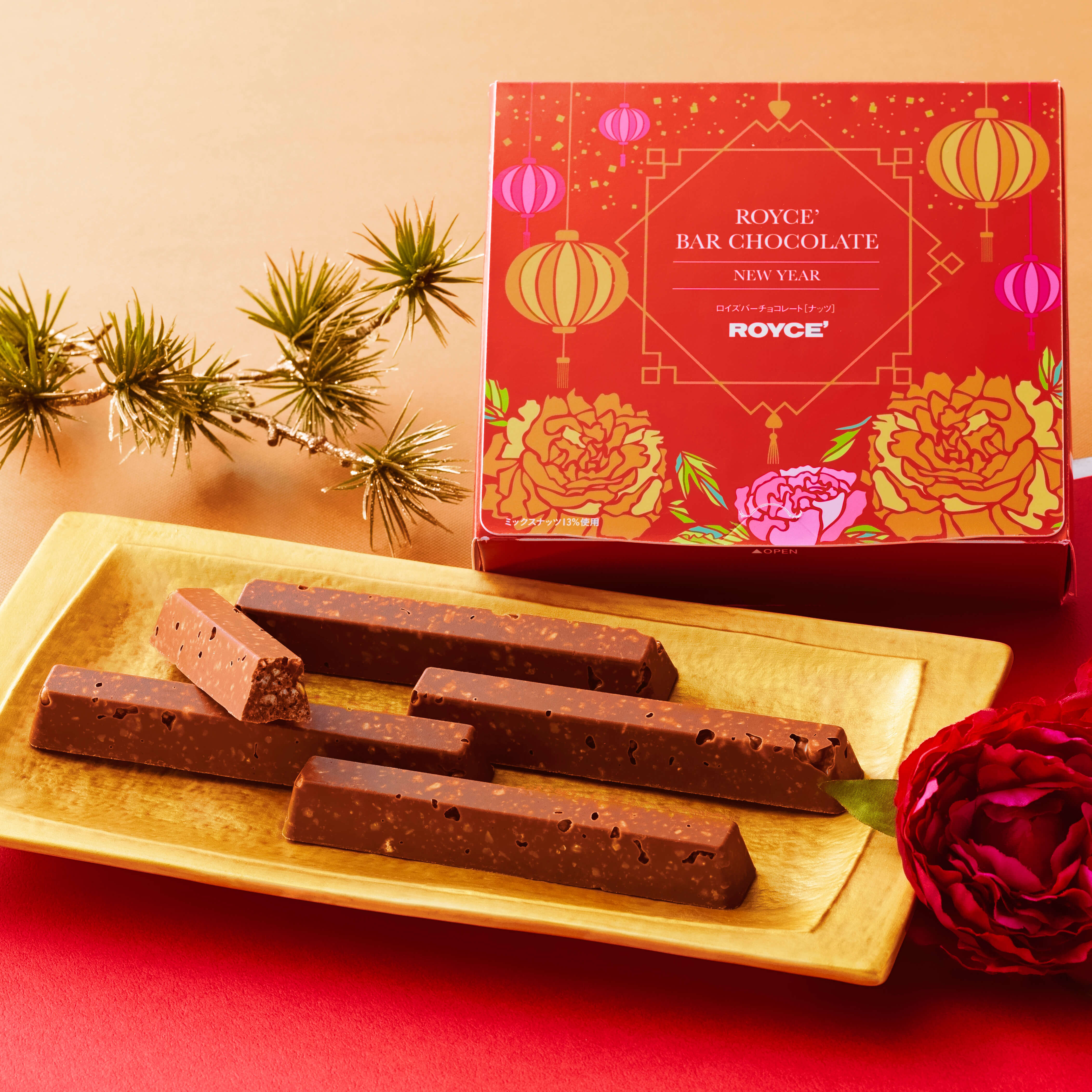 Image shows a red box with golden and pink prints of lanterns and flowers. Text on box says ROYCE' Bar Chocolate New Year ROYCE'. Accents include a golden tray with brown chocolate bars, two red flowers, and a golden twig with green leaves. Background is a combination of gold and red colors split in the middle.
