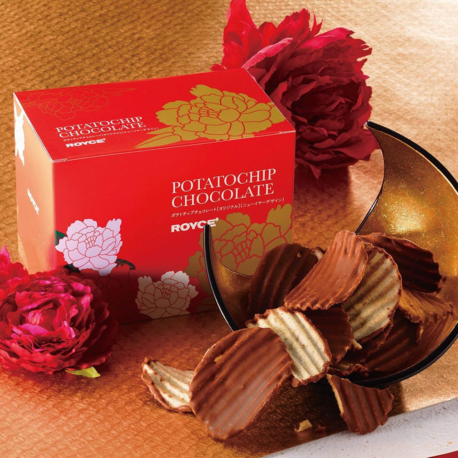 Image shows a red box with floral prints in gold, white, and pink. Overall text seen on box reads at Potatochip Chocolate ROYCE'. There is a gold crescent-shaped curved bowl with black edges. Accents include ridged potato chips in the colors brown and light yellow. Also in the picture are red flowers. Background is in gold with a hint of white in the bottom.