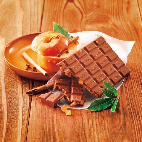 Image shows a brown chocolate bar on a crumpled white paper. Accents include a brown wooden bowl with a caramelized apple and some cinnamon rolls. Also seen are green leaves. Background is light brown.