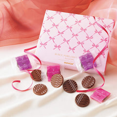 ROYCE' Chocolate - Pure Chocolate "Sweet & Milk" (Gift) - Image shows a pink box with ribbon prints and gold text saying ROYCE' Pure Chocolate Sweet & Milk ROYCE'. Accents include a pink ribbon, individually-wrapped chocolates in violet and pink, as well as brown chocolate discs with a waved texture. Background features a pink waved cloth on top and a silky white cloth below.