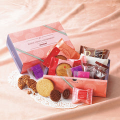 ROYCE' Chocolate - ROYCE' Sweet Assortment (17 Pcs) - Image shows two boxes with a nude-colored tablecloth. Box on top left is in the colors of blue and pink with various prints and text saying ROYCE' Sweet Assortment ROYCE'. Box in bottom middle right shows a blue and pink printed box that is filled with individually-wrapped confections in various shapes, sizes, and colors. Accent includes white circular table mats with cutouts.