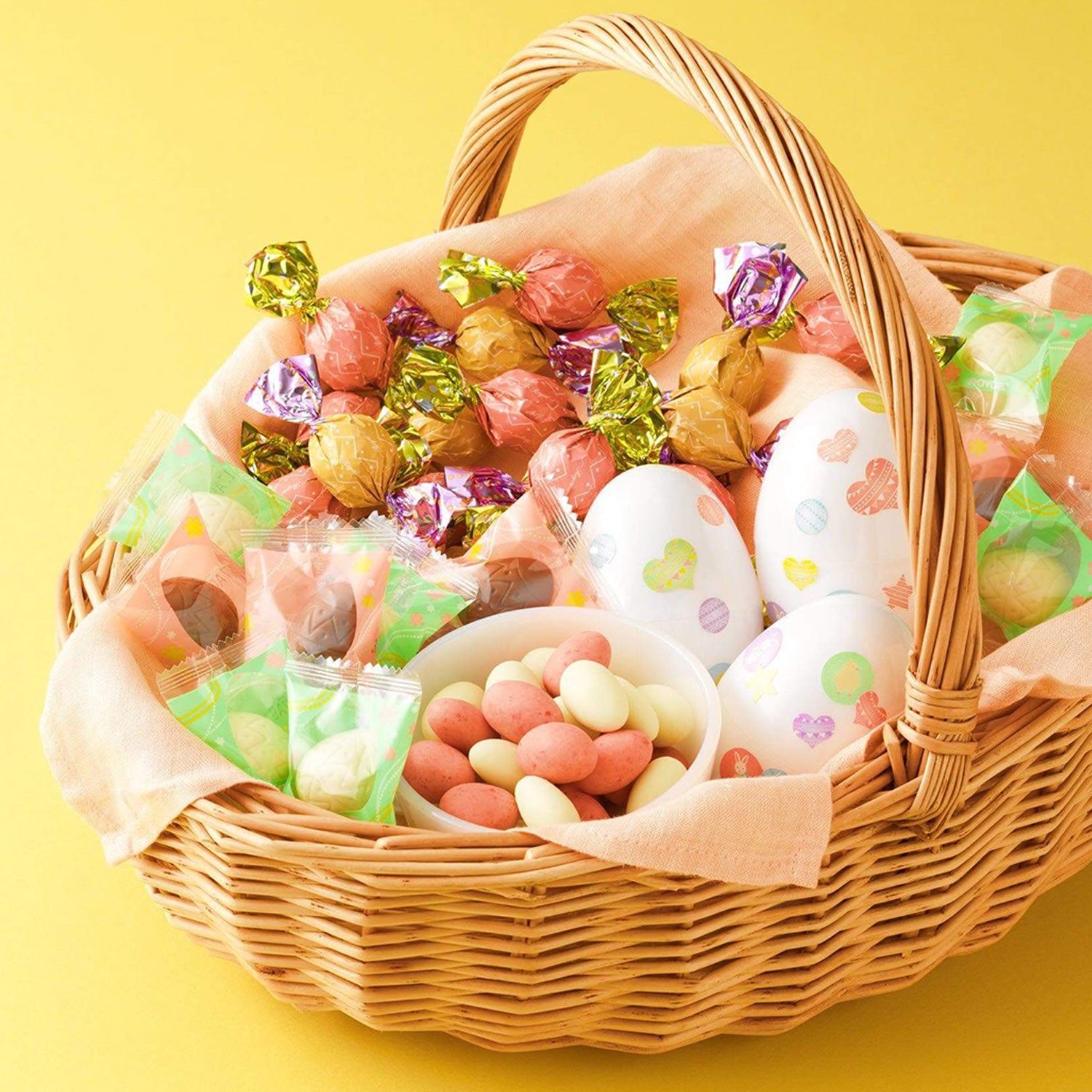 ROYCE' Chocolate - ROYCE' Chocolate Hunt Set - Image shows a brown wicker basket with peach-colored cloth filled with printed eggs and a variation of confections in different colors and shapes. Background is in yellow.