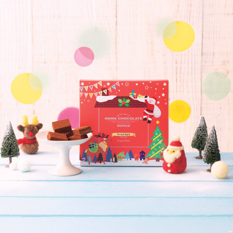 Image shows a red box in the middle with illustrations of a reindeer, Santa Claus, trees, plants, a roof with chimney, and stars. Text says Merry Christmas ROYCE' Quality Nama Chocolate ROYCE' Mild Milk Liquor Free. Accents include chocolate blocks on a white bowl and charms of trees, snowballs, a reindeer, and Santa Claus. Background is light brown wood with yellow and pink circles. Surface is wood painted in blue.
