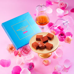Image shows a box of chocolates in blue with some chocolates on a plate and some wine glasses and petals.