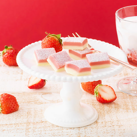 Image shows pink and white chocolate blocks on a white elevated plate. Accents include strawberries and a glass of milk with strawberry. Background is in red.