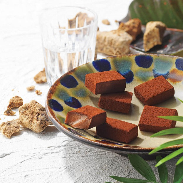 ROYCE' Chocolate - ROYCE' Ishigakijima Nama Chocolate "Kokutoh" - Image shows brown blocks of chocolate on a printed plate. Accents include green leaves, brown sugar blocks, and a glass of water.