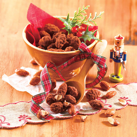 Roasted Almond Chocolate - Image shows brown clusters of chocolate-covered almonds inside a brown bowl with red paper, red berries, and green leaves. There are also chocolate-coated almond clusters on a brown table and a wooden spoon with a printed red ribbon with stripes of green, white, and black. Accents include a white crumpled paper, a wooden toy soldier, and a flesh-colored table cloth with red trimmings. Background is in light brown.
