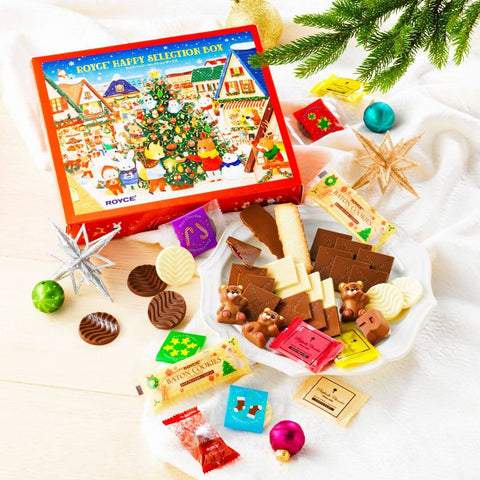 ROYCE' Happy Selection Box - Image shows a printed box on top left with illustrations of animals, houses, and a decorated tree. Text says ROYCE' Happy Selection Box. Accents include chocolates and confections, a white plate, decorative orbs and stars, a white tablecloth, and green mistletoe.