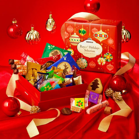 ROYCE' Holidays Selection (32 Pcs) - Image shows a printed red box with illustrations of decoration balls and lamps. Text says ROYCE' Holidays Selection. Also seen is an open red box with varying individually-wrapped confections. Accents include a decorative orbs, gold acorns, gold ribbon, and loose confections. Background is in red.