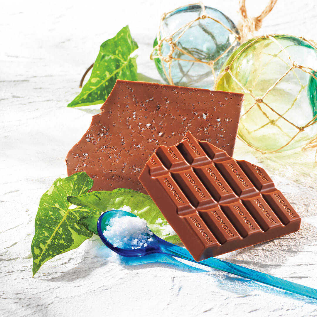 ROYCE' Chocolate - ROYCE' Ishigakijima Ishigaki no Shio Chocolate - Image shows brown chocolate bars on a white surface with green leaves. Accents include a blue spoon with salt and blue and yellow glass orbs with golden nets.