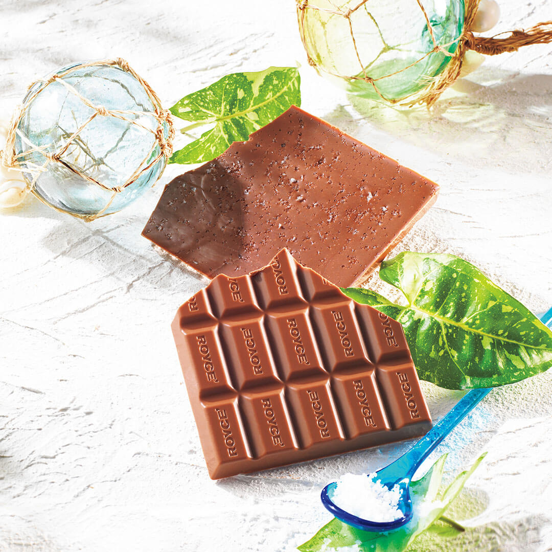 ROYCE' Chocolate - ROYCE' Ishigakijima Ishigaki no Shio Chocolate - Image shows brown chocolate bars on a white surface with green leaves. Accents include a blue spoon with salt and blue and yellow glass orbs with golden nets.