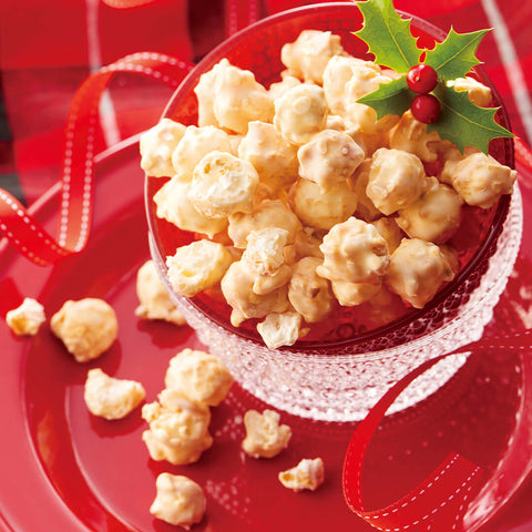 Image shows popcorn coated with white chocolate in a red bowl on a clear glass and red plate. Accents include a red ribbon and green leaves with red berries. Background is in red.