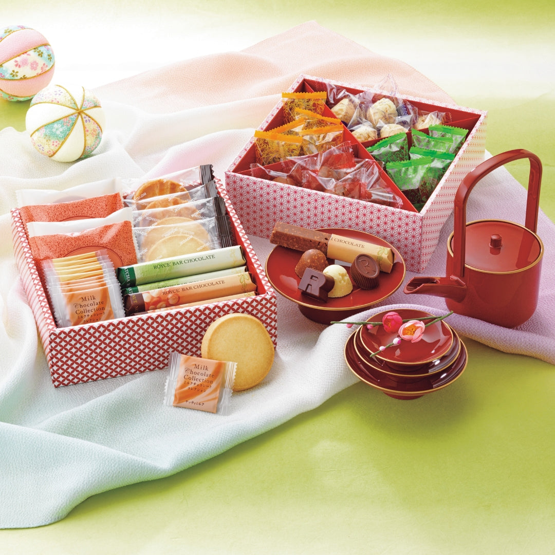 ROYCE' Sweets Osechi - Image shows open printed boxes with individually-wrapped confections inside. Accents include red plates and a red tea pot. Also seen are decorative balls and loose confections.