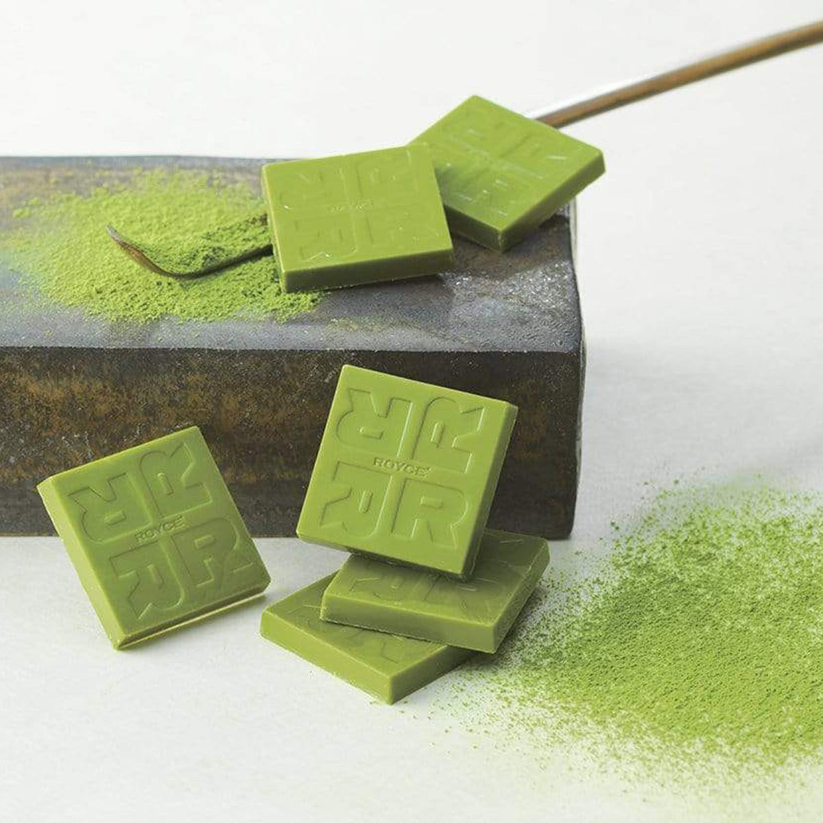 ROYCE' Chocolate - Matcha Chocolate - Image shows green chocolate squares etched with "R" letters and the word "ROYCE'". Accents include a brown wooden stick, green tea powder, and a gray stone platform. Background is white.